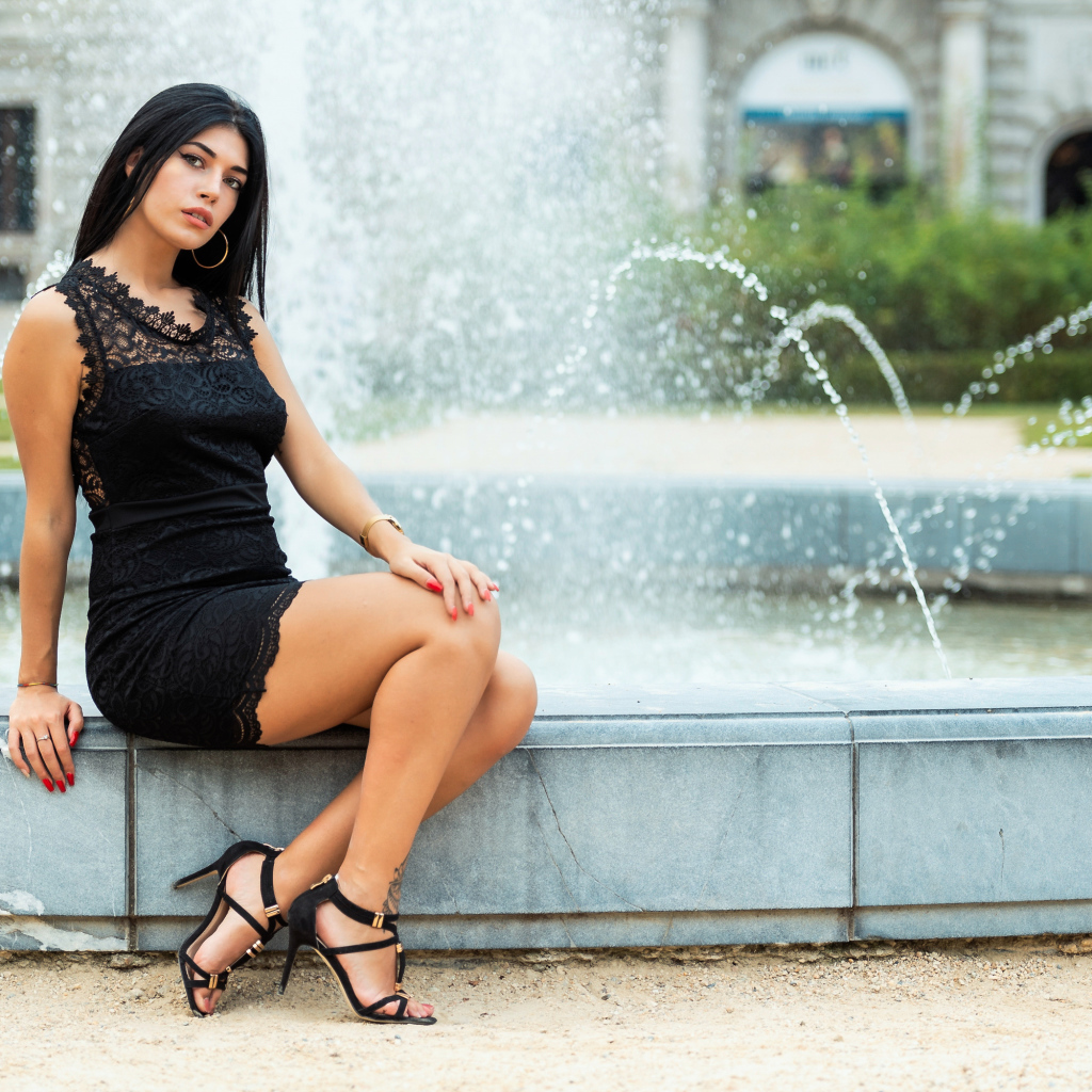 Красивая брюнетка в черном платье сидит у фонтана