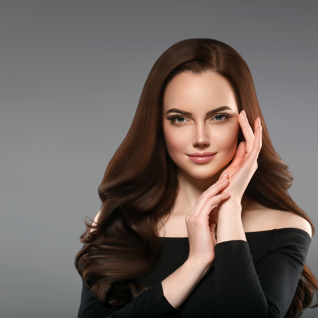 Красивая нежная длинноволосая шатенка  с руками у лица на сером фоне