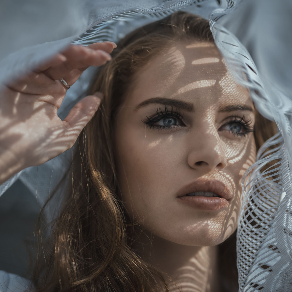 Голубоглазая девушка смотрит в окно с тюлью