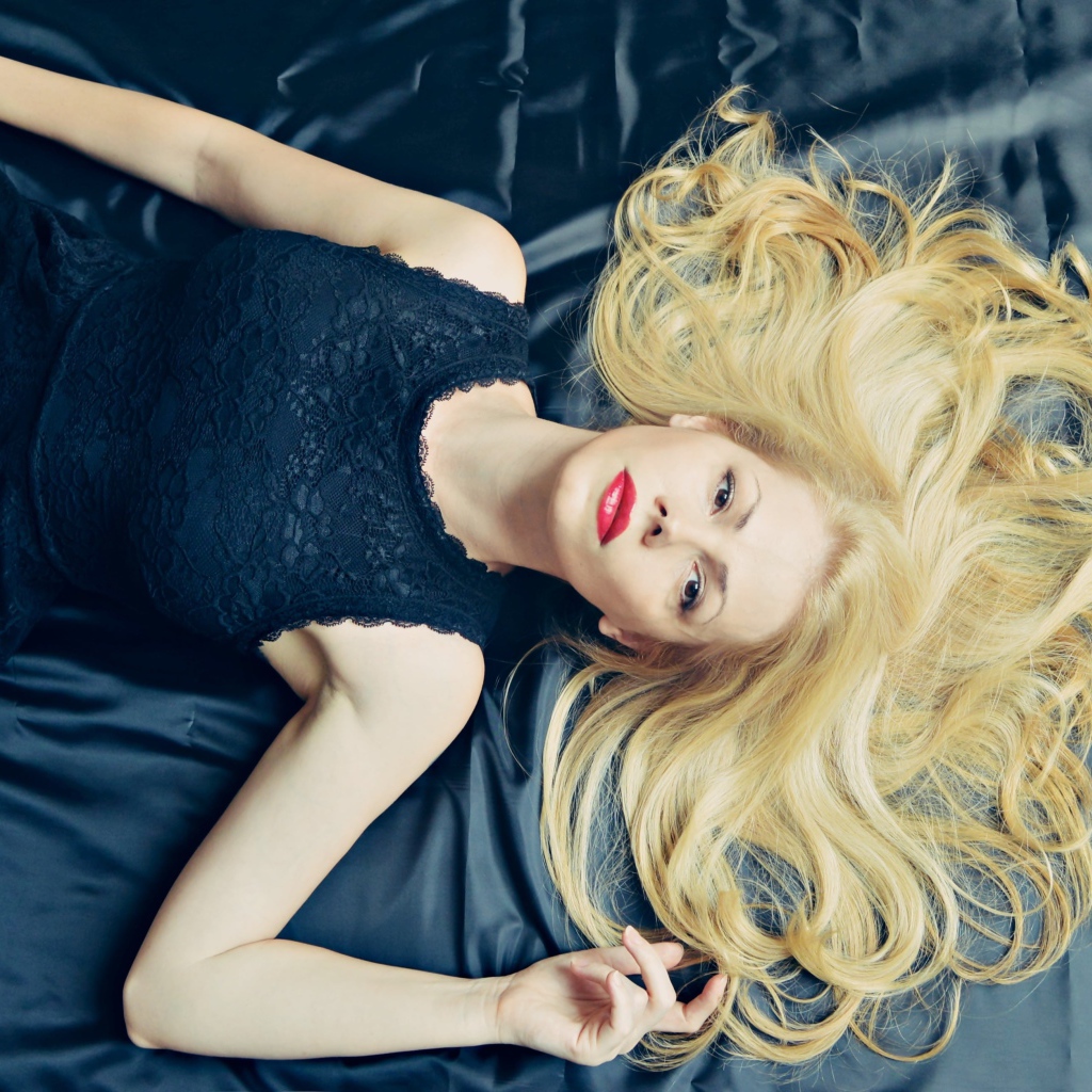 Длинноволосая блондинка в черном платье лежит на кровати