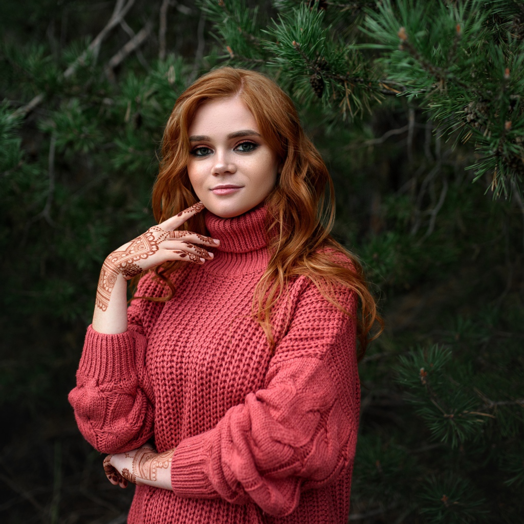 Рыжеволосая девушка в розовом свитере стоит у ели