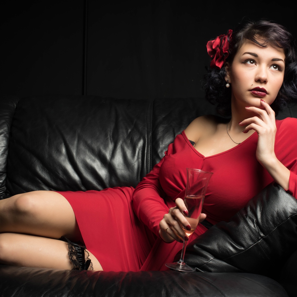 Девушка в красном платье с бокалом в руке лежит на кожаном диване