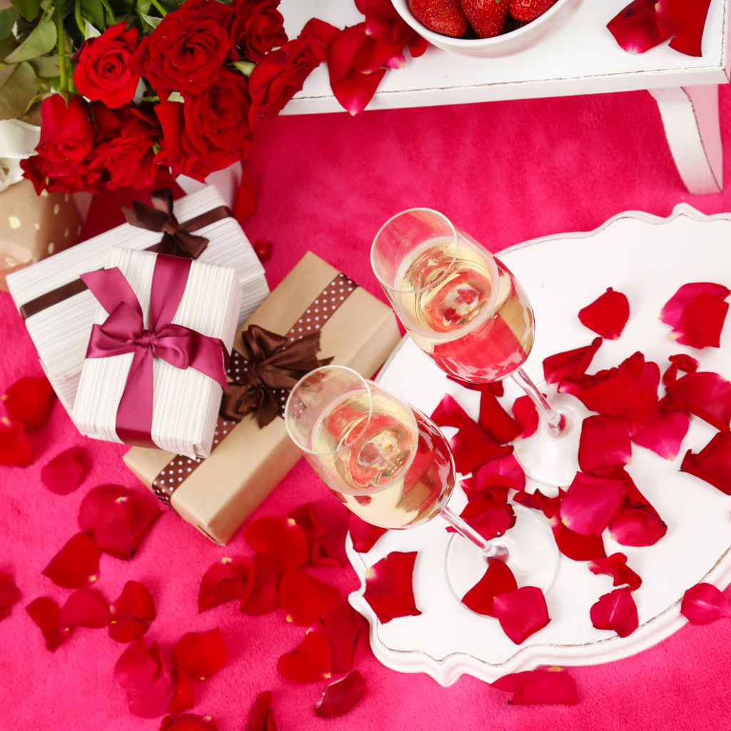 Два фужера с шампанским на красном покрывале с лепестками, подарками и букетом роз для любимой