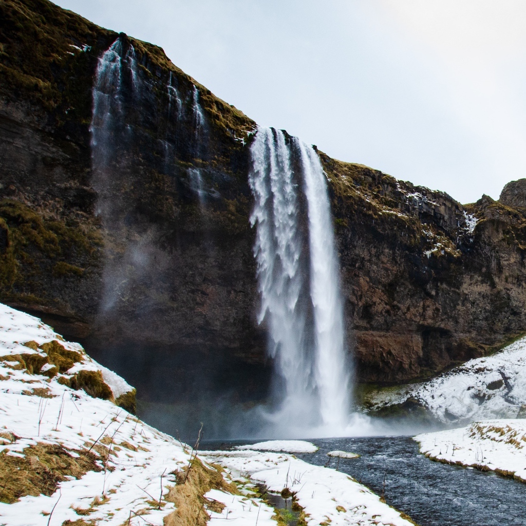 Водопад стекает со скалы в реку с покрытыми снегом берегами