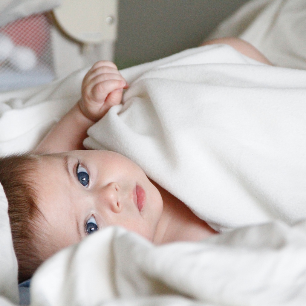 Маленький грудной голубоглазый ребенок лежит под белым покрывалом 