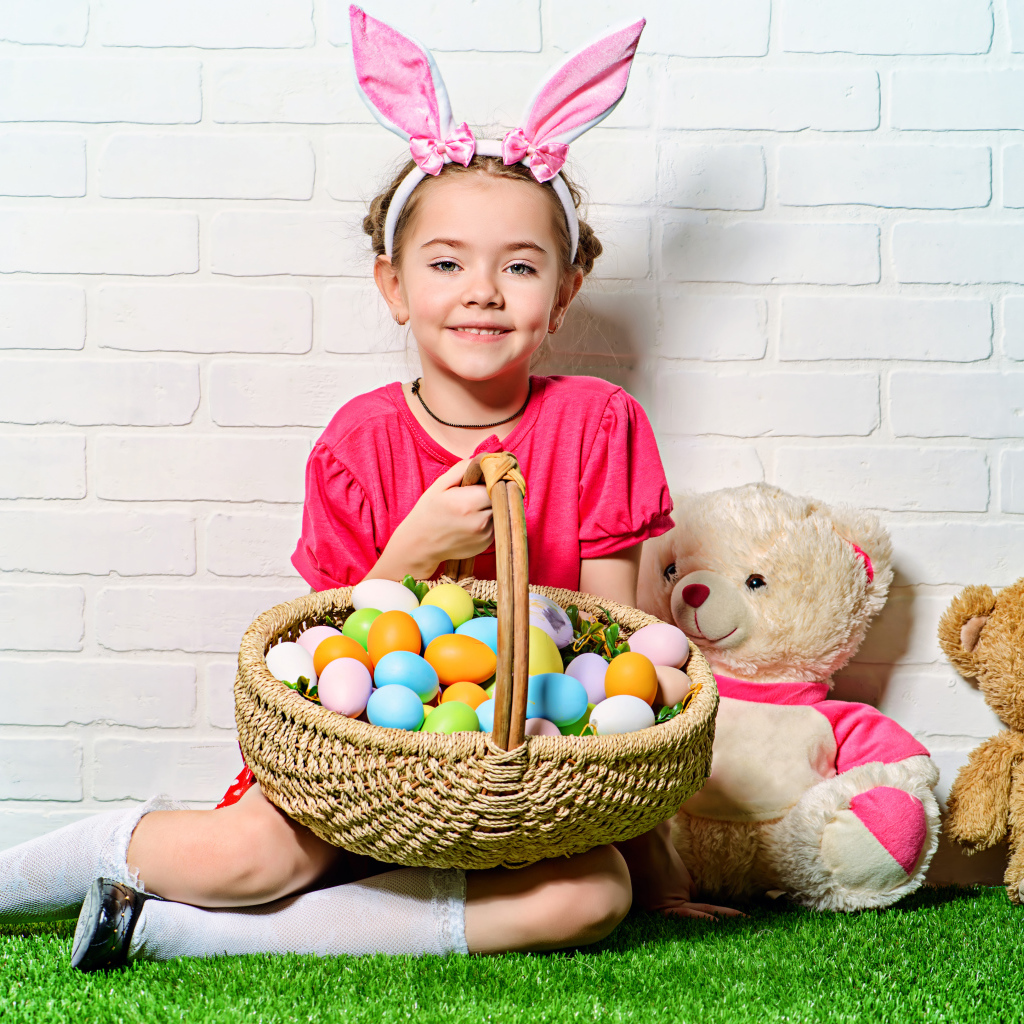 Улыбающаяся девочка сидит у стены с корзиной пасхальных яиц и игрушками
