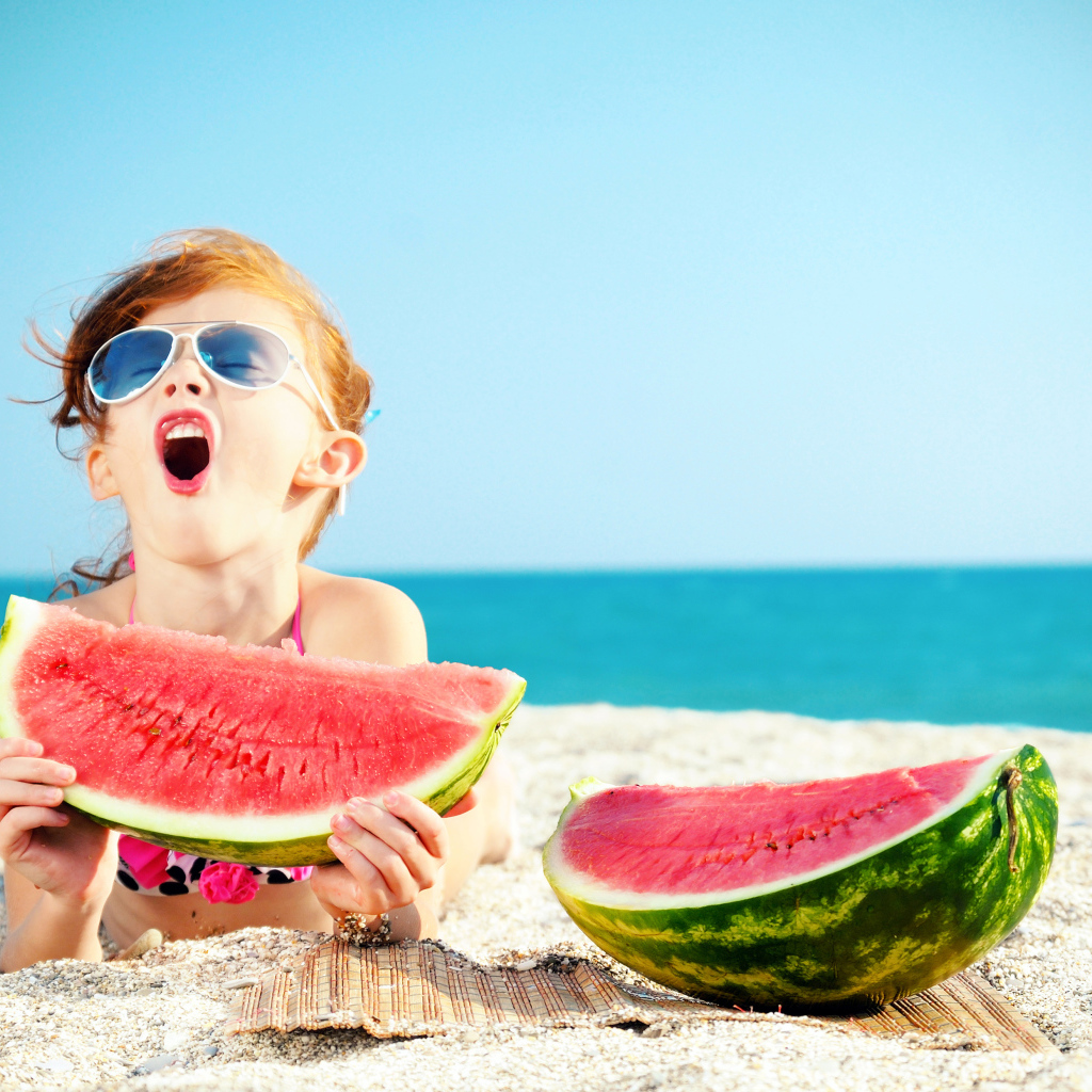 Маленькая девочка ест арбуз на песке у моря