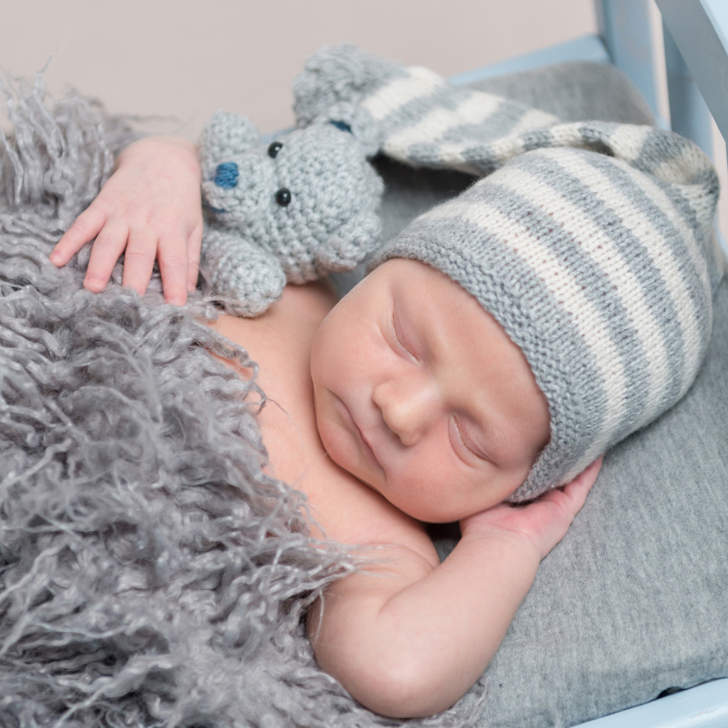 Младенец в вязаной шапке спит с игрушкой под меховым одеялом