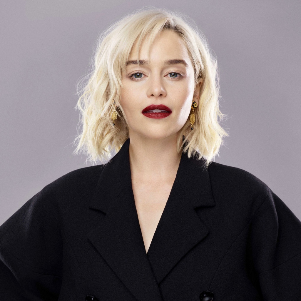 Яркая блондинка, актриса Эмилия Кларк в черном пальто на сером фоне