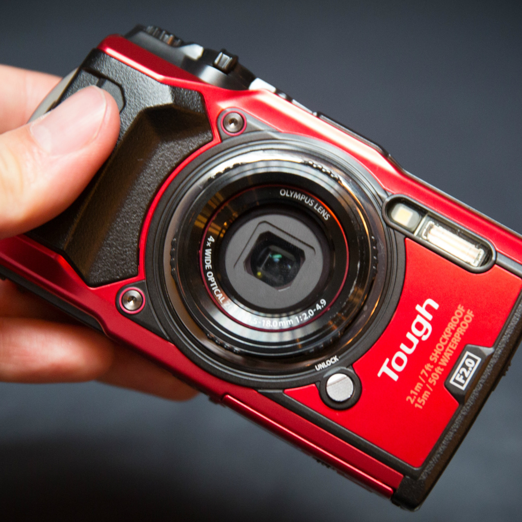 Красный фотоаппарат Olympus Tough TG-5 в руке на сером фоне