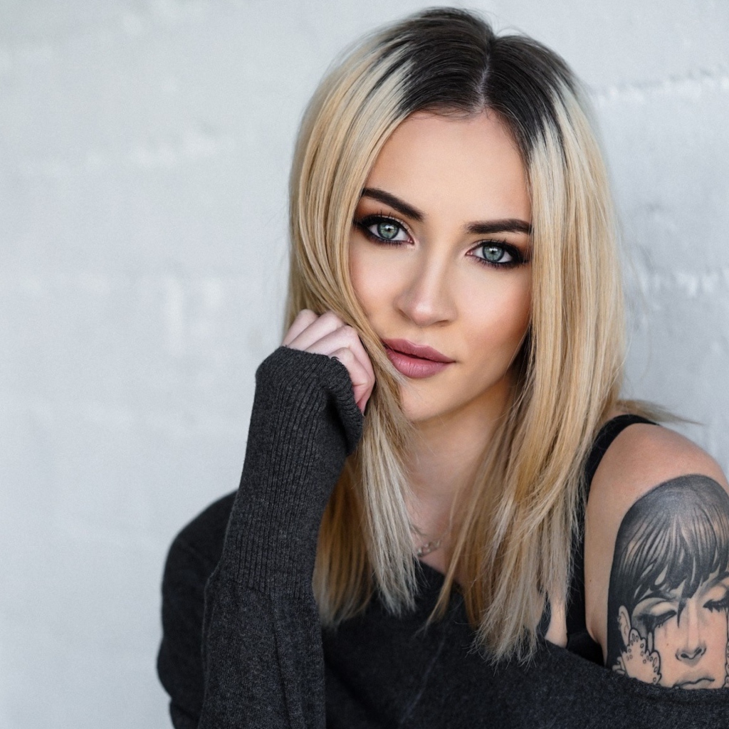 Стильная блондинка с татуировкой на плече на фоне стены