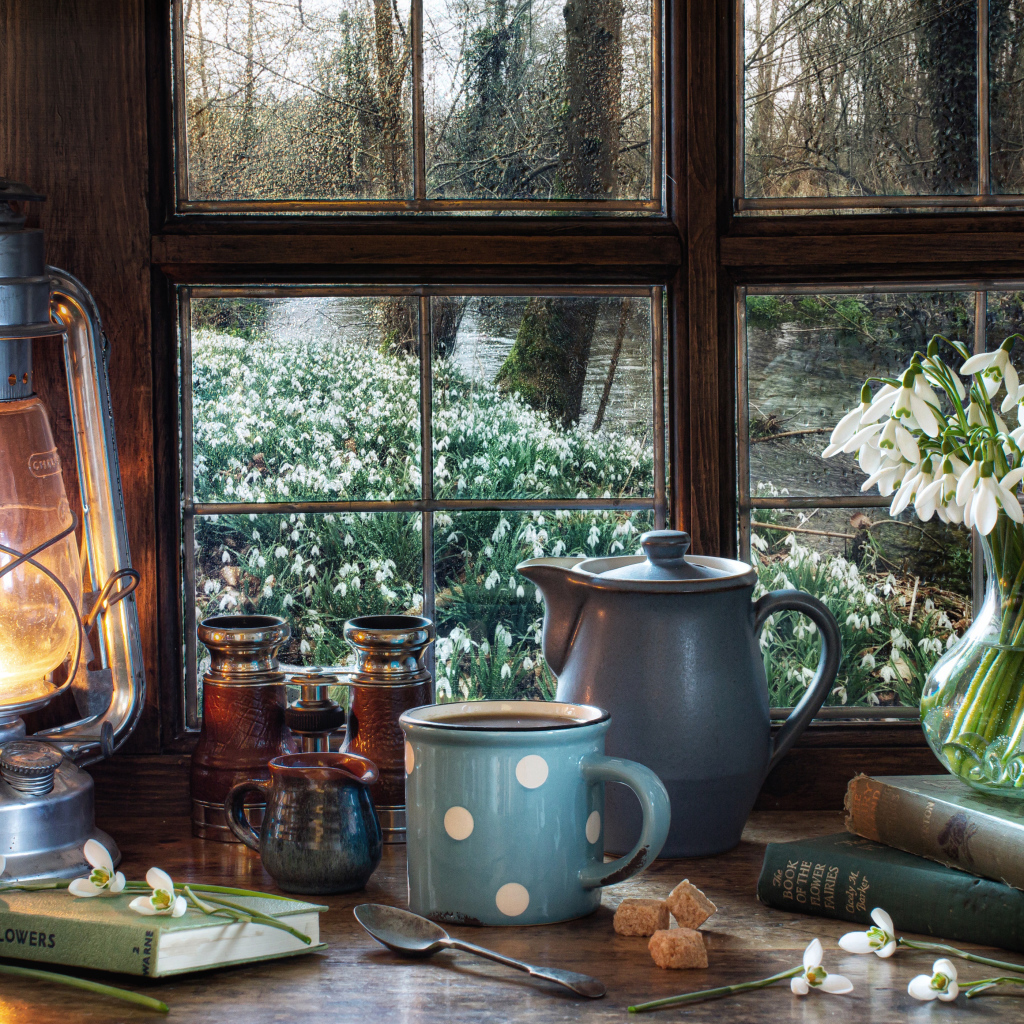 Керосиновая лампа на столе с кружкой, книгами и букетом подснежников у окна