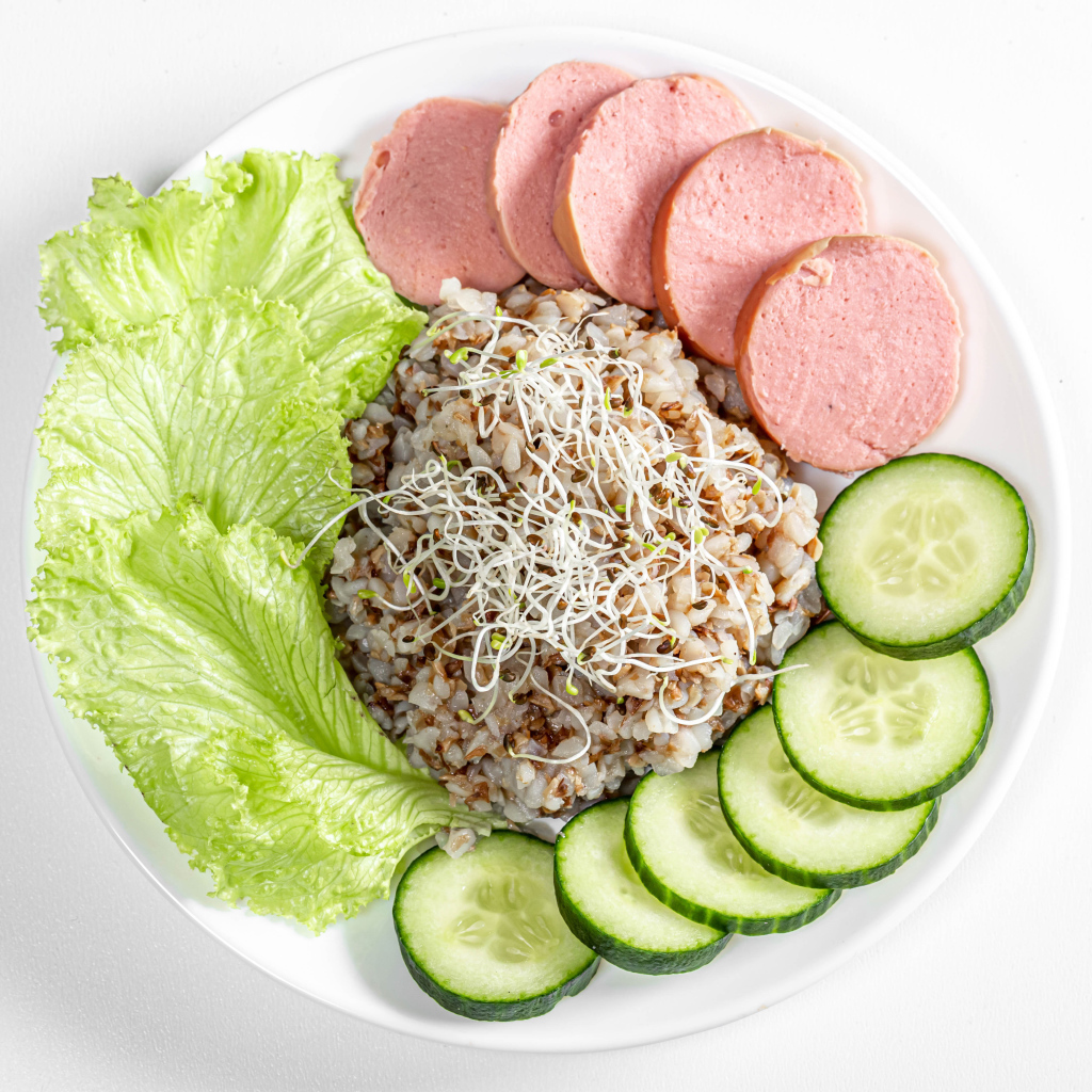 Гречка на тарелке с огурцом, колбасой и листьями салата на белом фоне