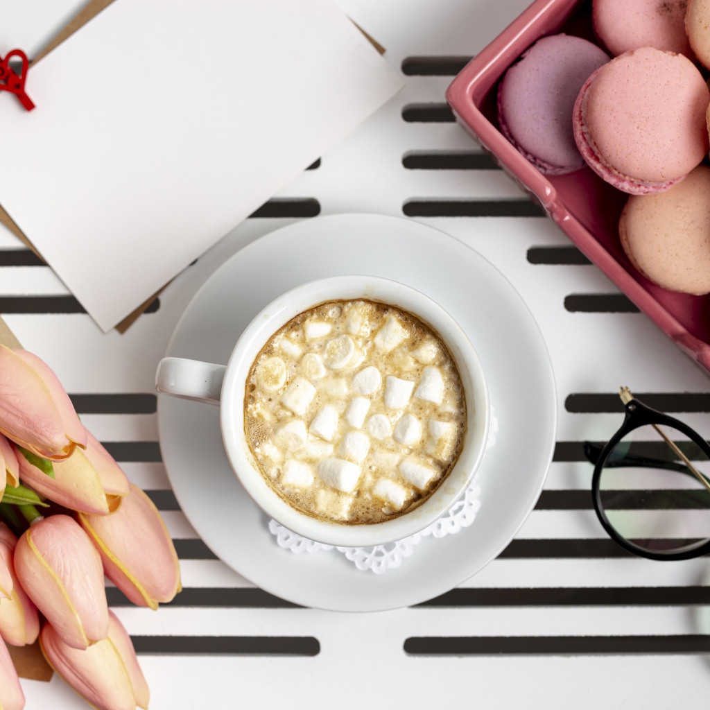 Чашка какао с маршмеллоу на столе с букетом тюльпанов, десертом макарун и очками