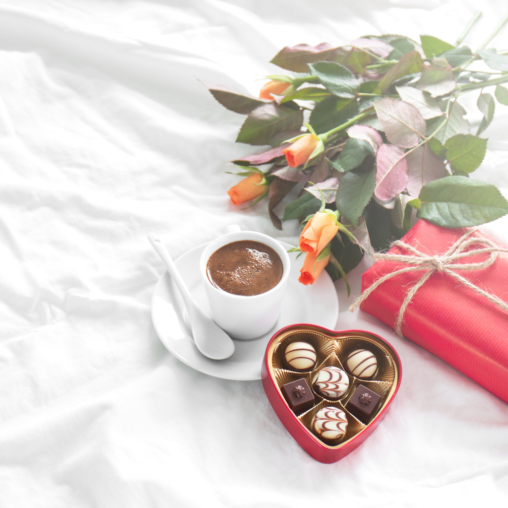Чашка кофе на белой кровати с конфетами, подарком и букетом роз