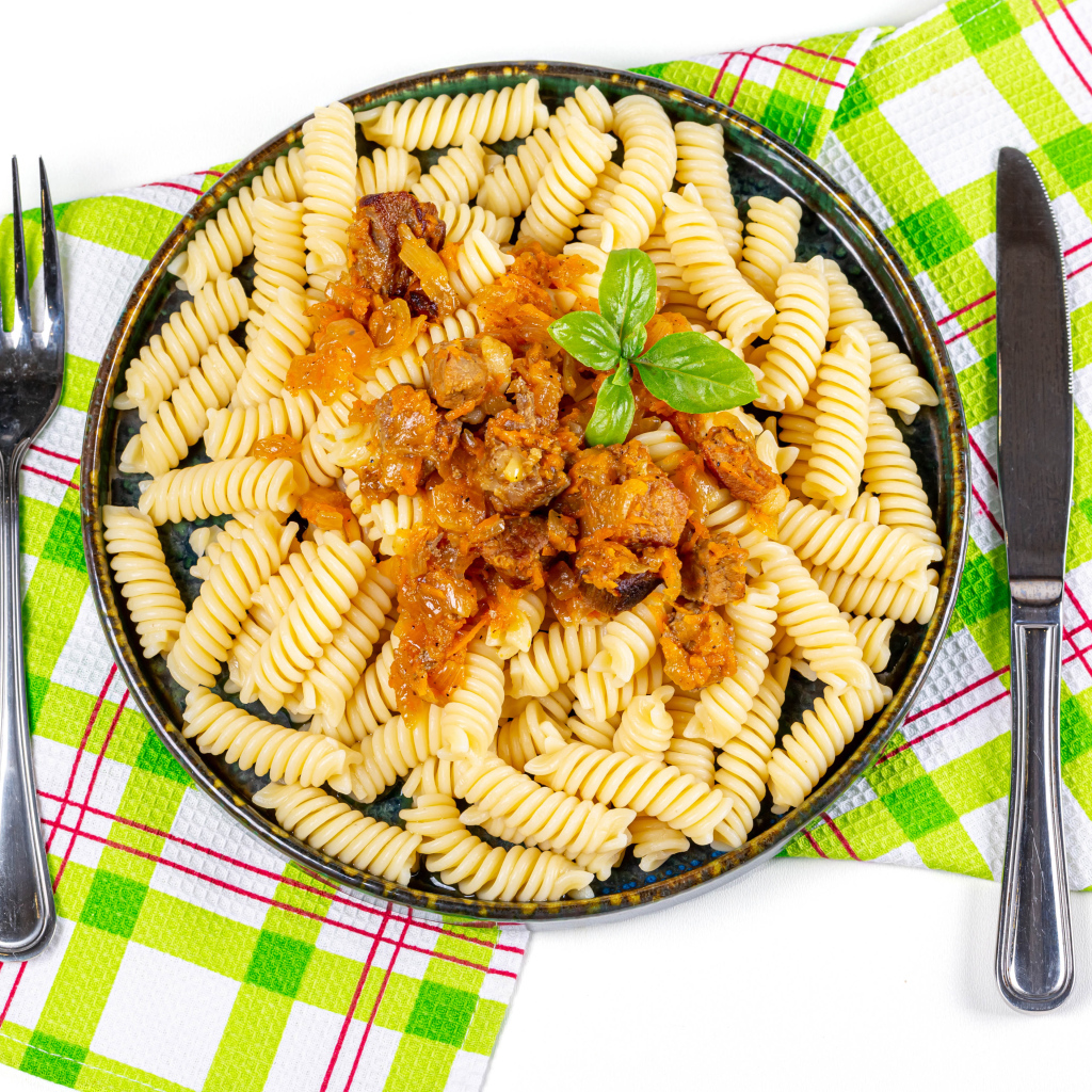 Большая тарелка макарон с мясом на столе со столовыми приборами