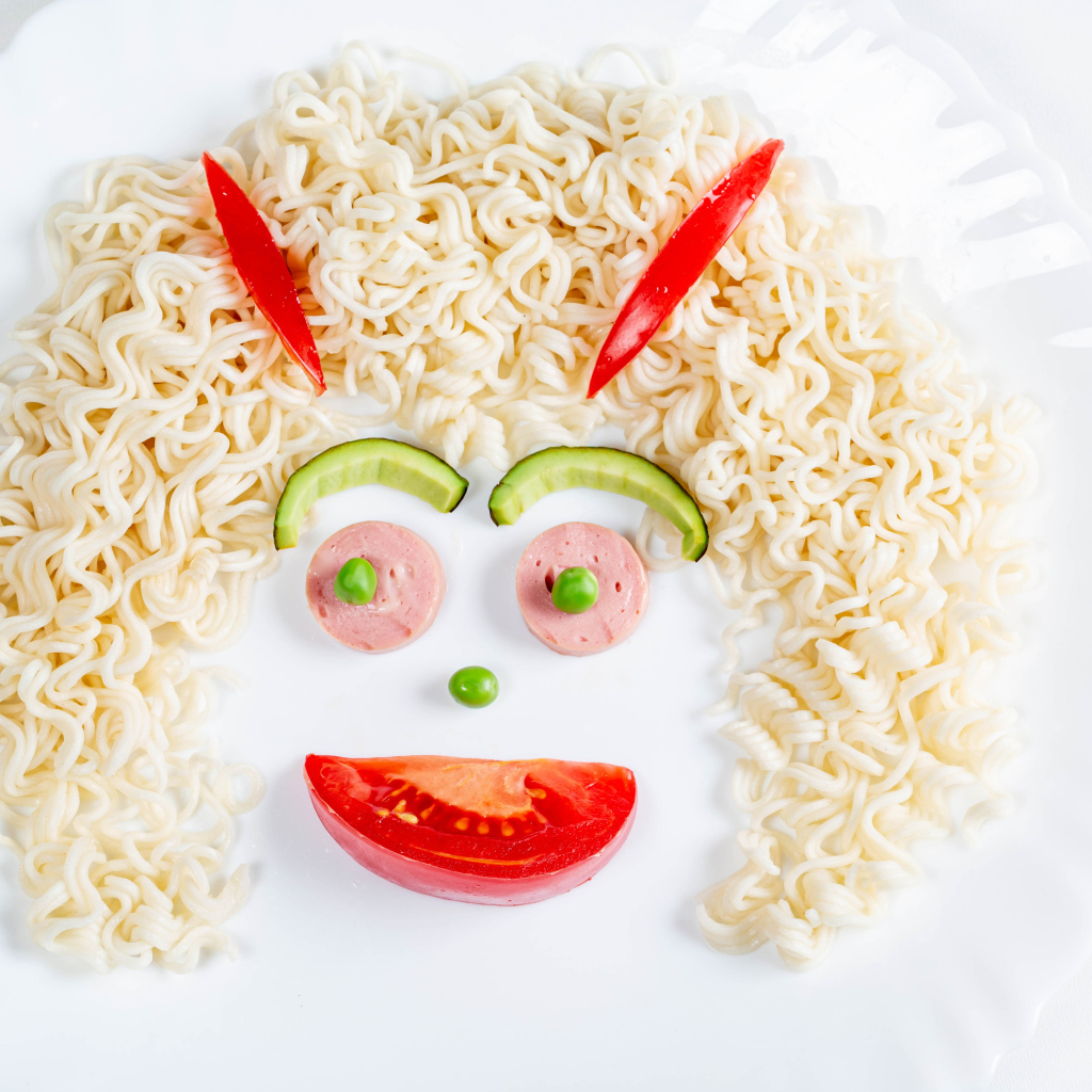 Лицо из вермишели с овощами и колбасой на белой тарелке 