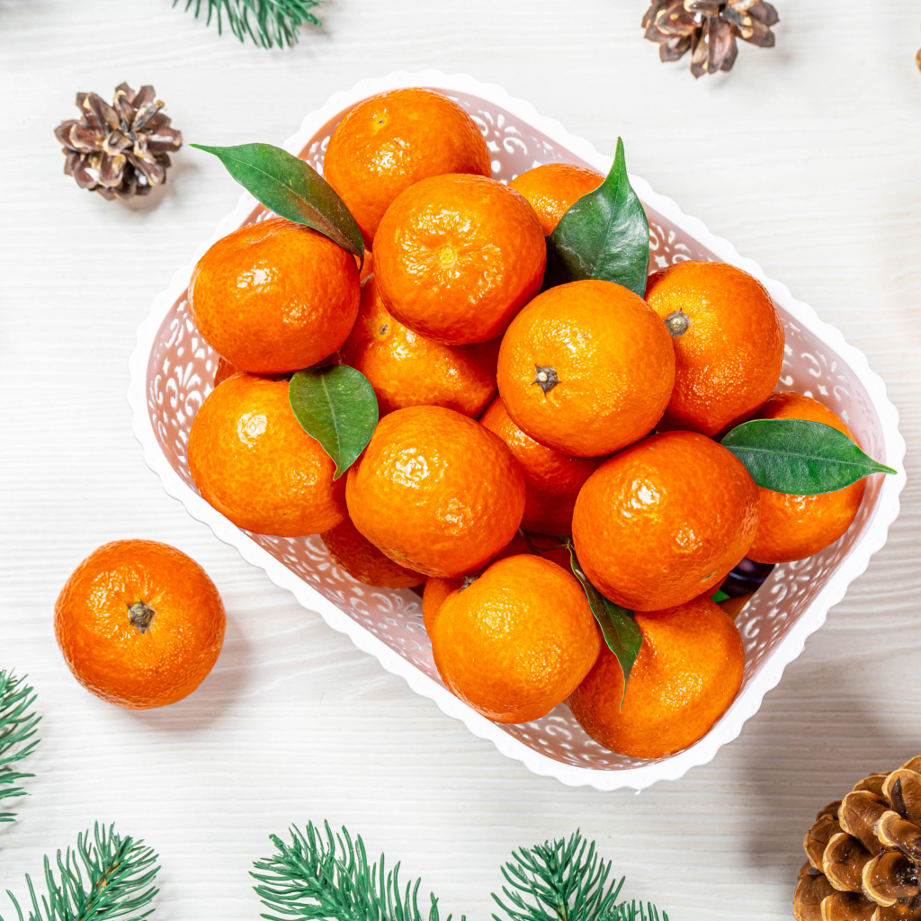 Много спелых мандаринов в белой корзинке на столе с шишками