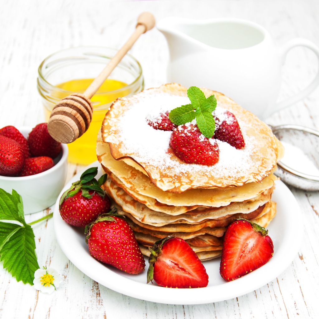 Тонкие блины с сахарной пудрой и ягодами клубники на столе с молоком и медом