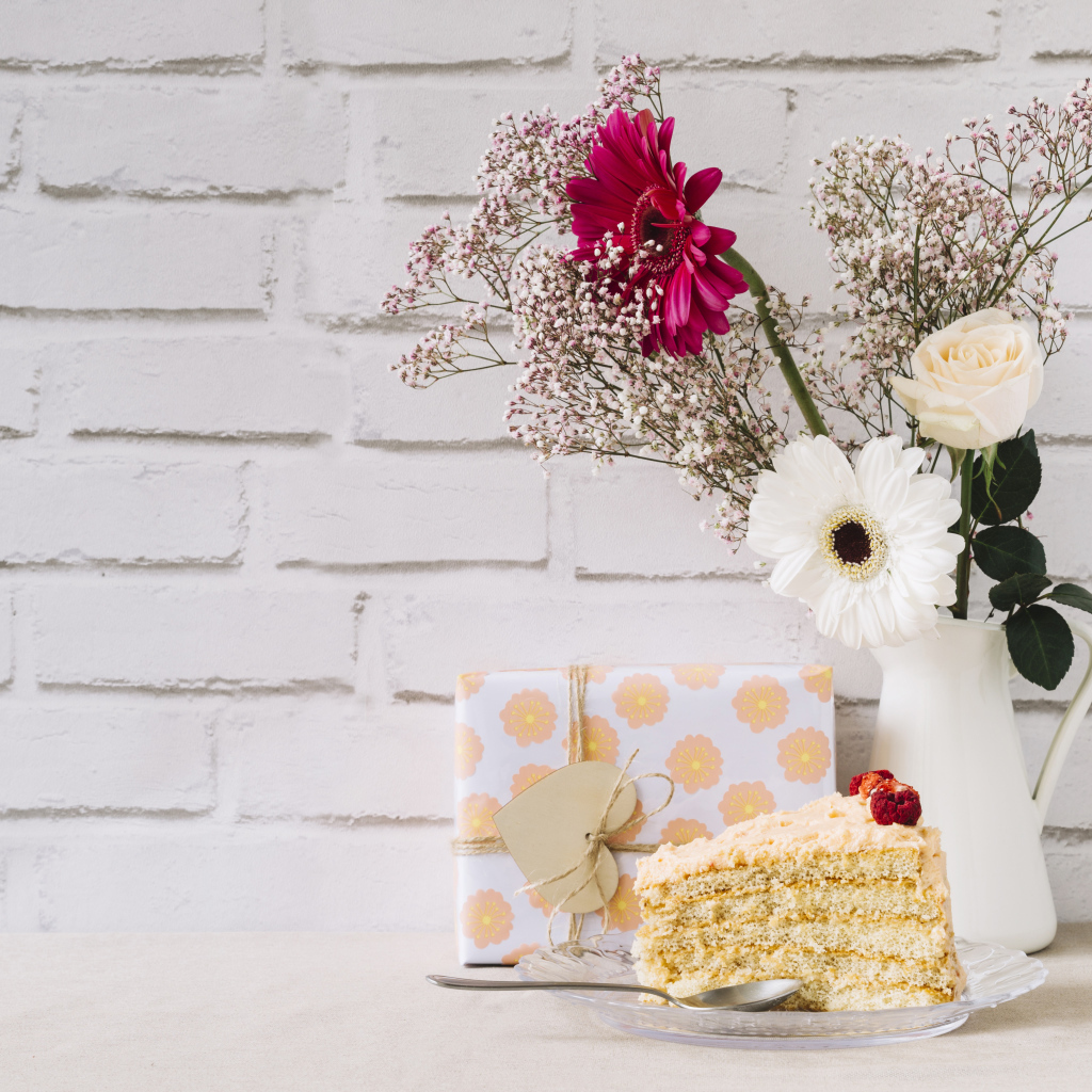 Кусок торта, подарок и букет на столе на фоне стены 