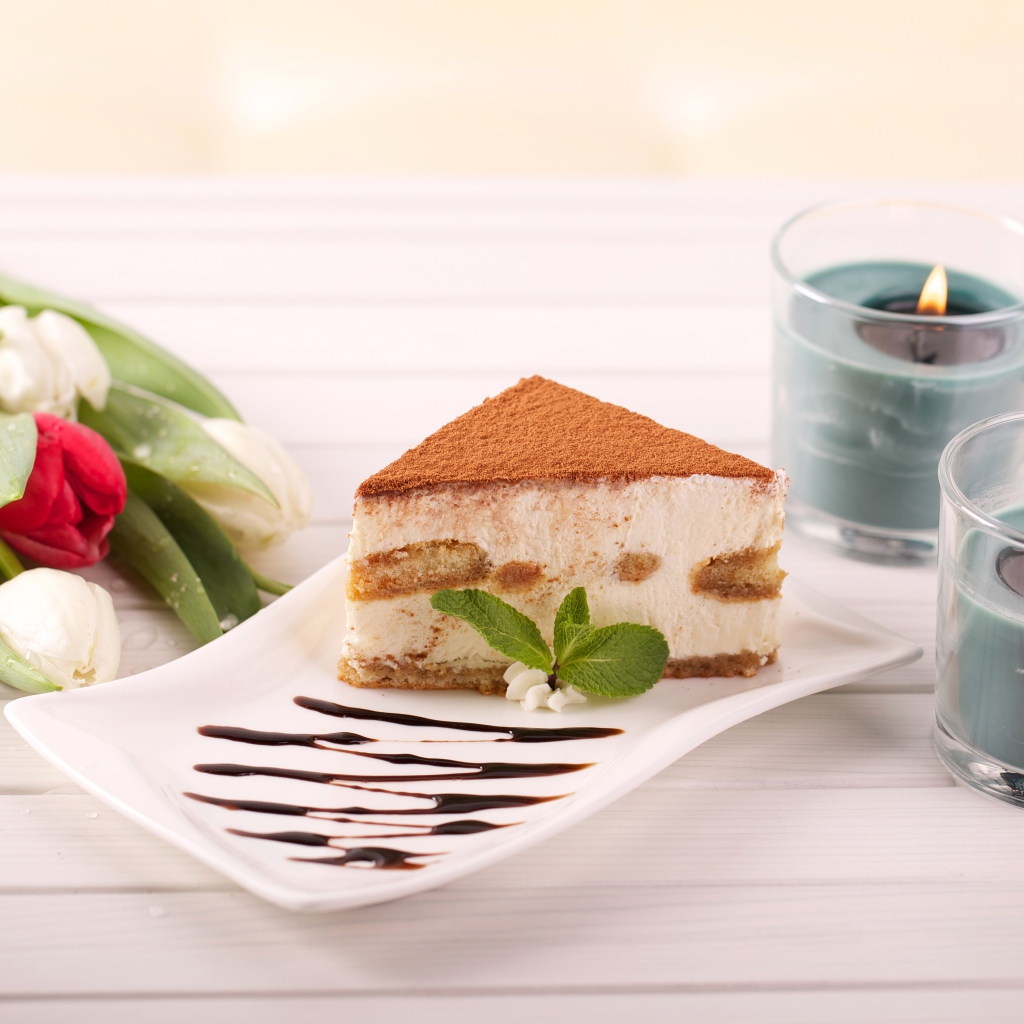 Кусок тирамису на столе с зажженными свечами и букетом тюльпанов