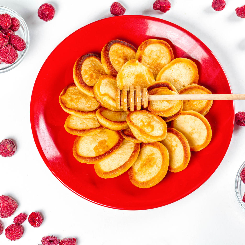 Оладьи на красной с медом на красной тарелке на столе с ягодами малины