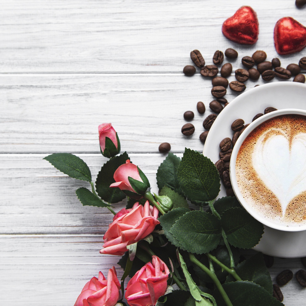 Чашка кофе с рисунком на пене на столе с букетом роз, зернами и конфетами