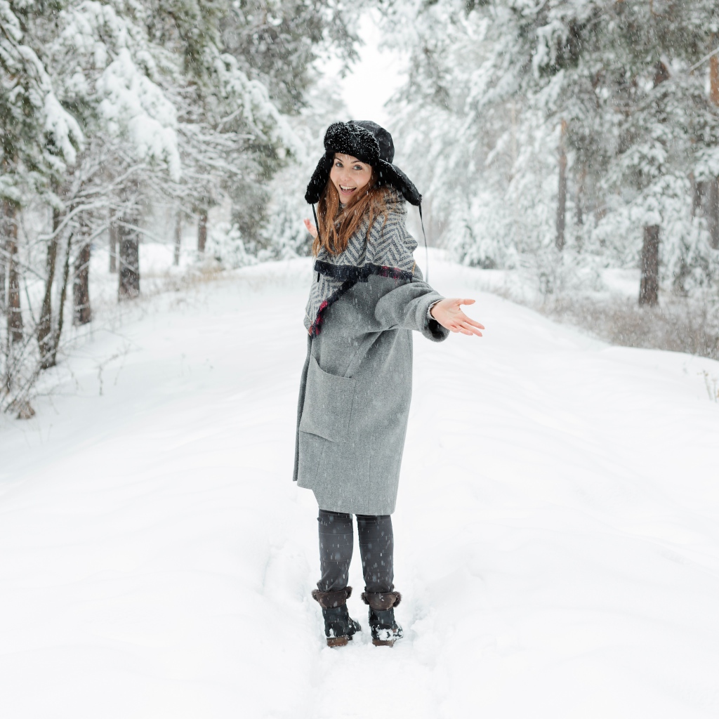 Девушка в пальто и теплой шапке гуляет в заснеженном лесу 