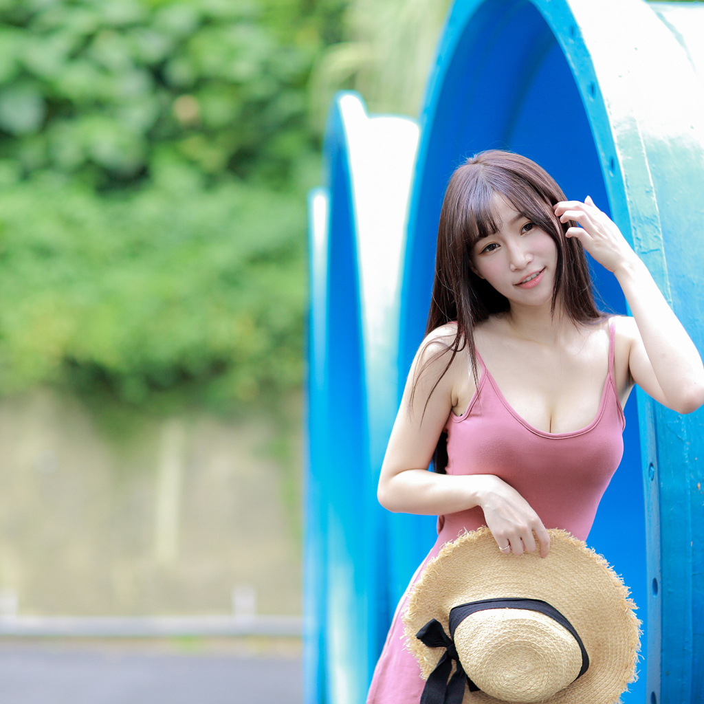 Стройная азиатка в платье со шляпой в руке стоит у синих  труб 