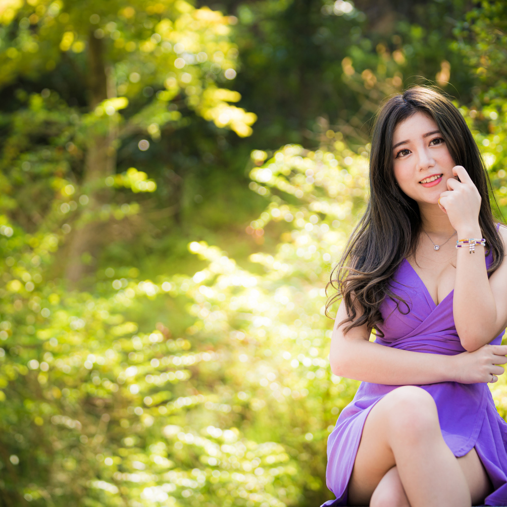 Улыбающаяся девушка азиатка в фиолетовом платье сидит в парке 
