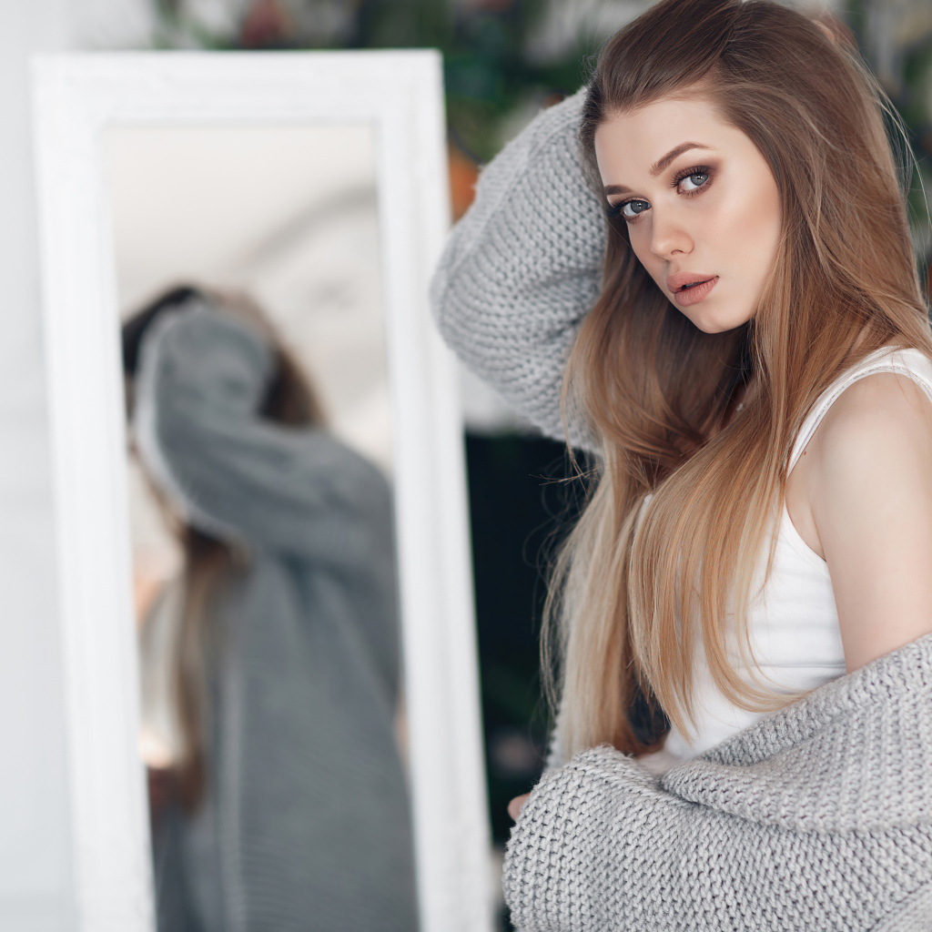 Молодая девушка в серой кофте стоит у зеркала 