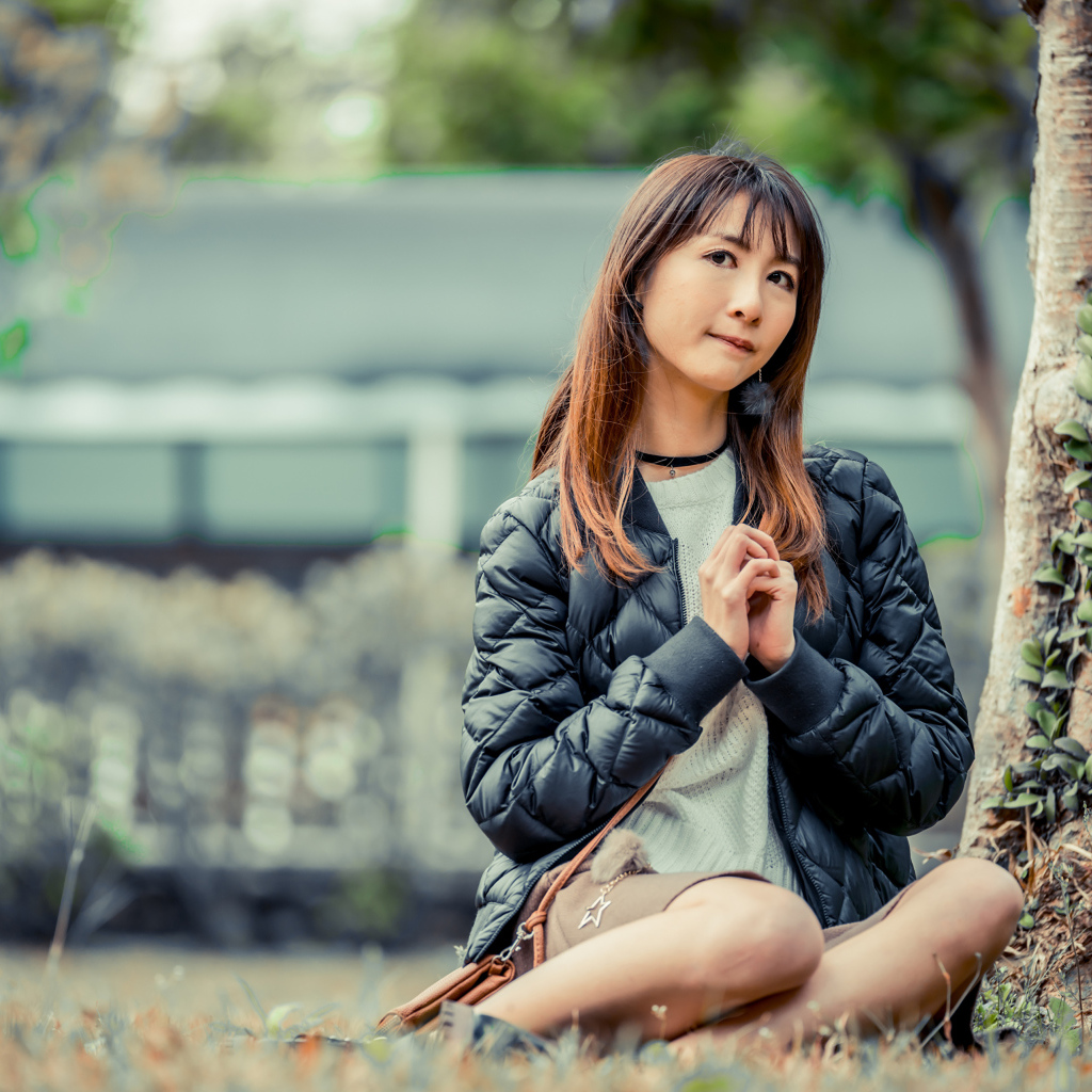 Девушка азиатка в черной куртке сидит под деревом