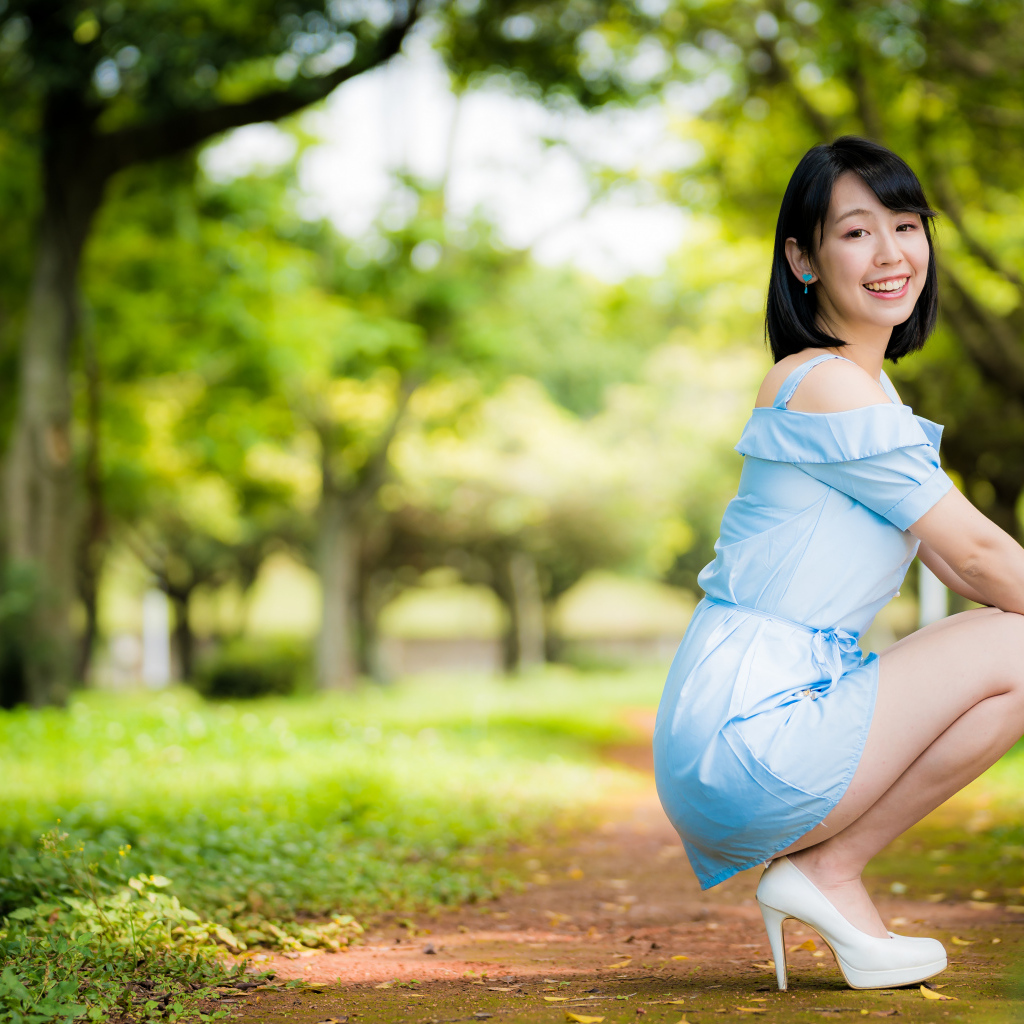 Девушка азиатка в голубом платье сидит на корточках в парке