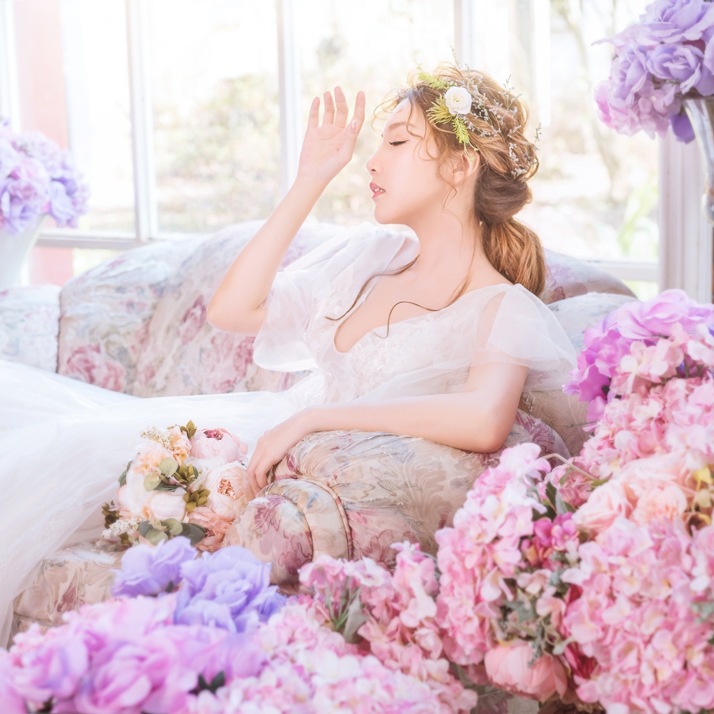 Девушка азиатка в белом платье сидит на диване в комнате с цветами