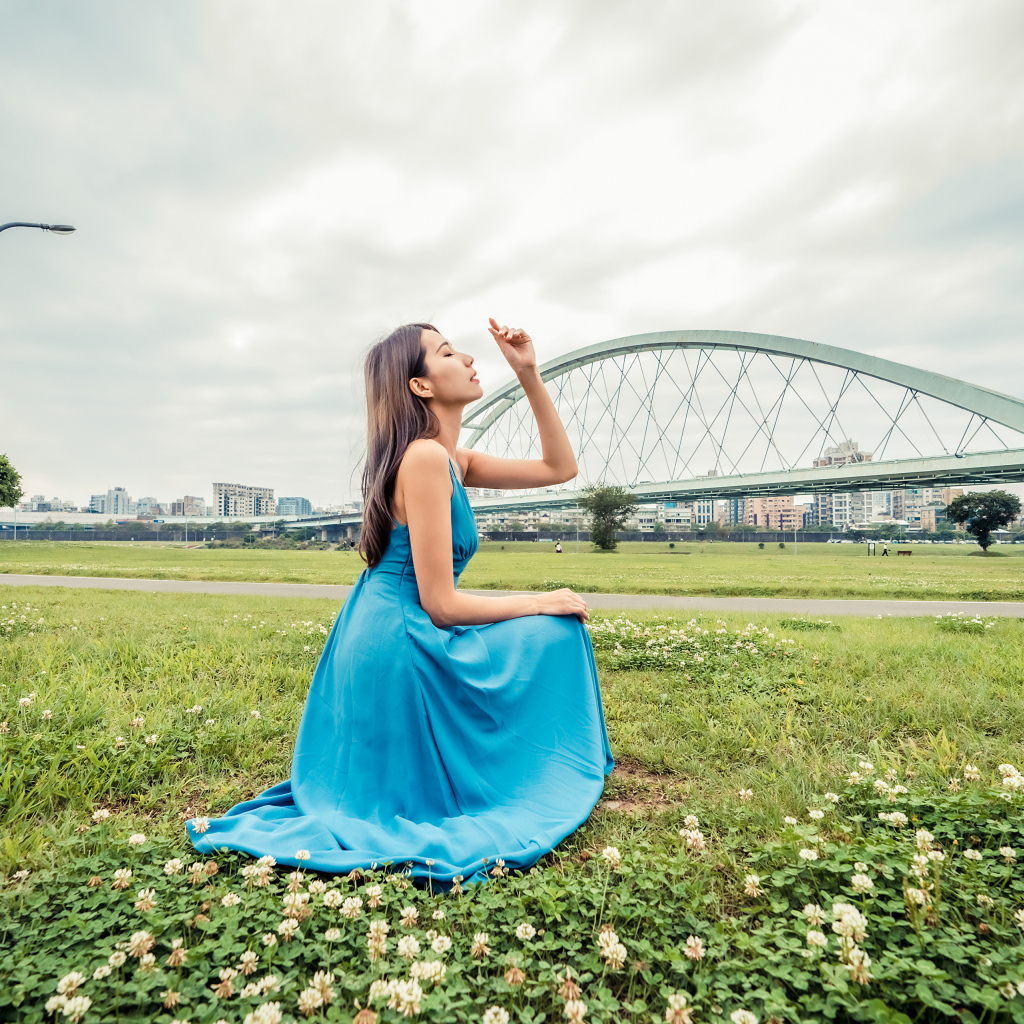 Красивая девушка азиатка в голубом платье  сидит на траве на фоне моста