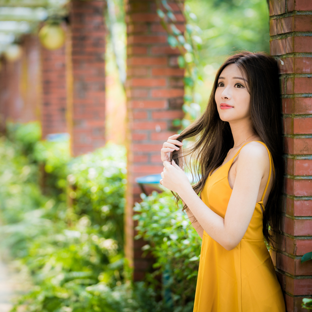 Красивая азиатка в желтом платье стоит у кирпичной колонны 