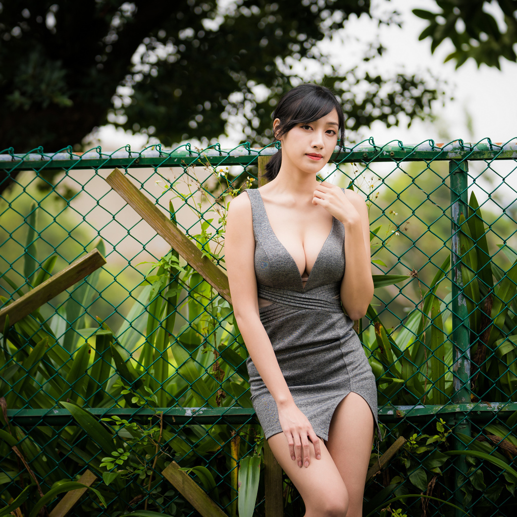 Красивая азиатка в сером платье стоит у зеленой сетки 