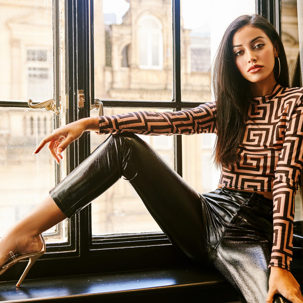 Красивая брюнетка в черных брюках сидит на окне 