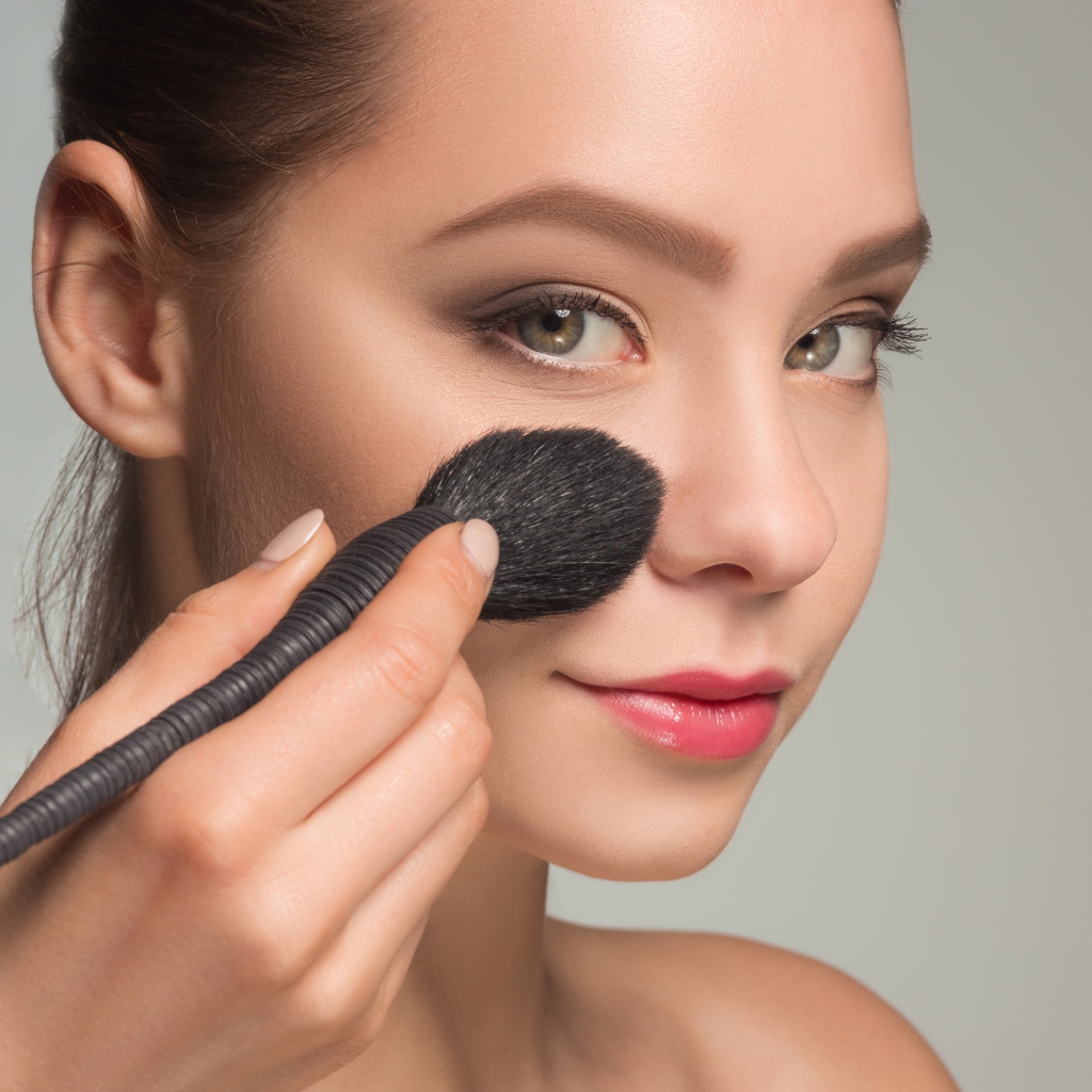 Красивой девушке наносят макияж на лицо кистью 