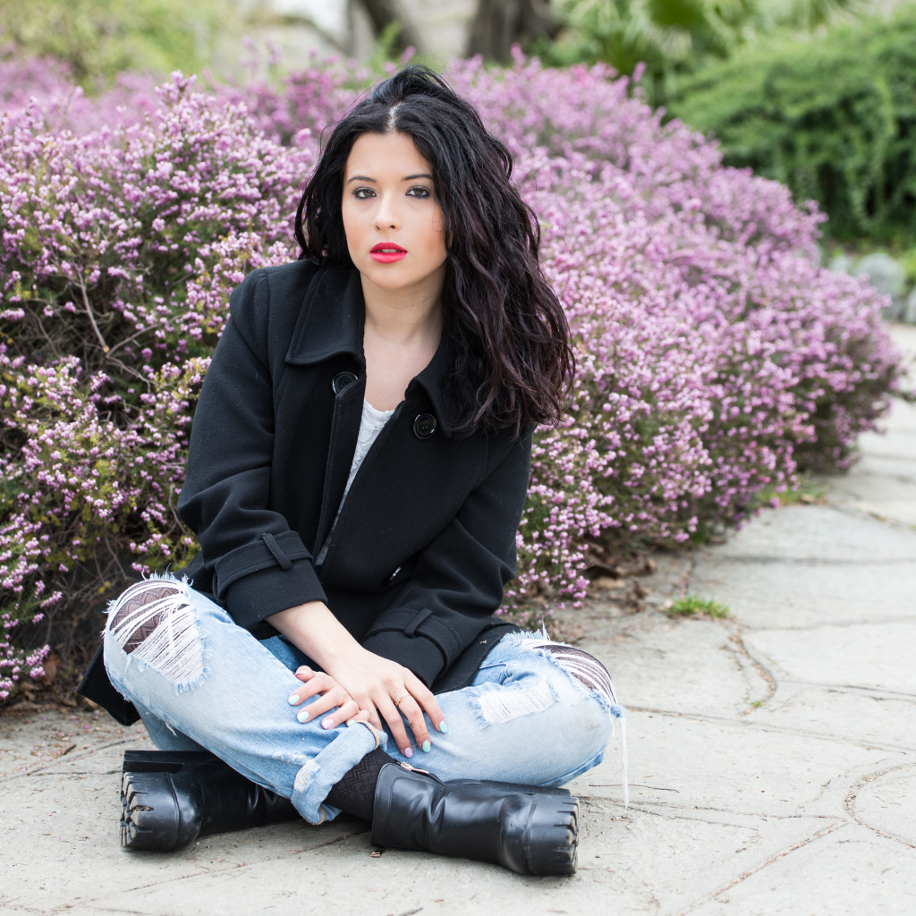 Красивая девушка в черном пальто сидит на земле у кустов