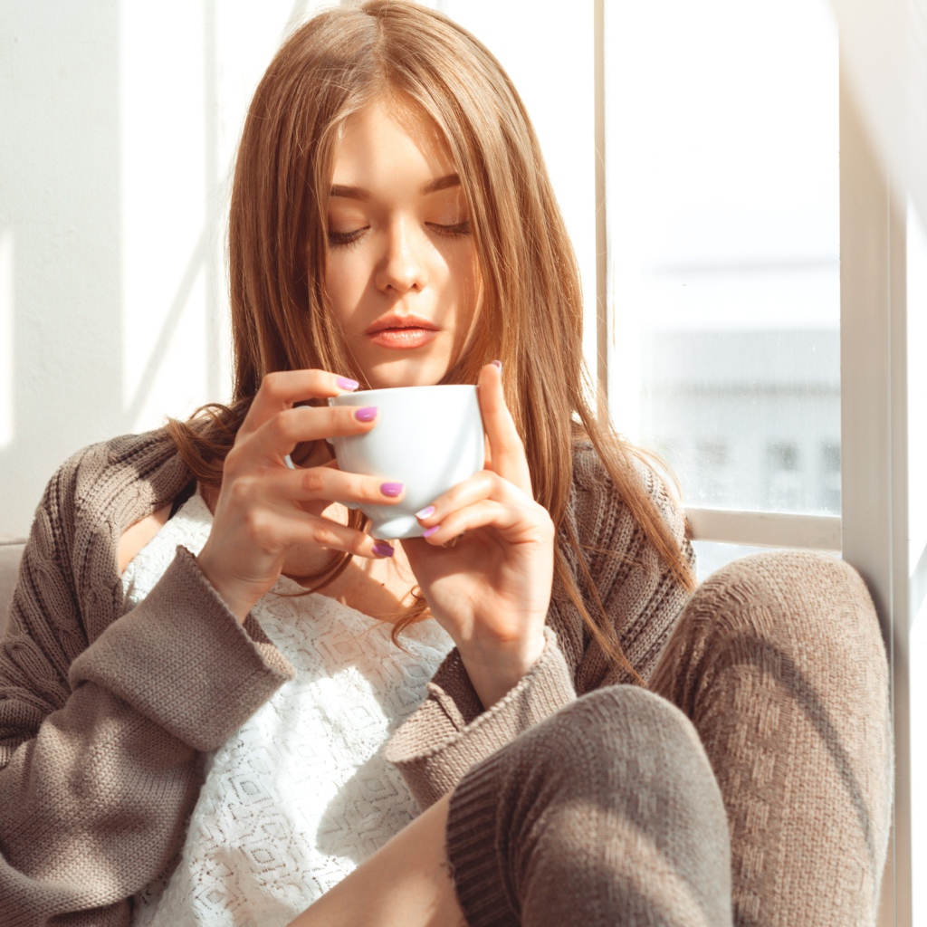 Красивая девушка в свитере пьет кофе у окна