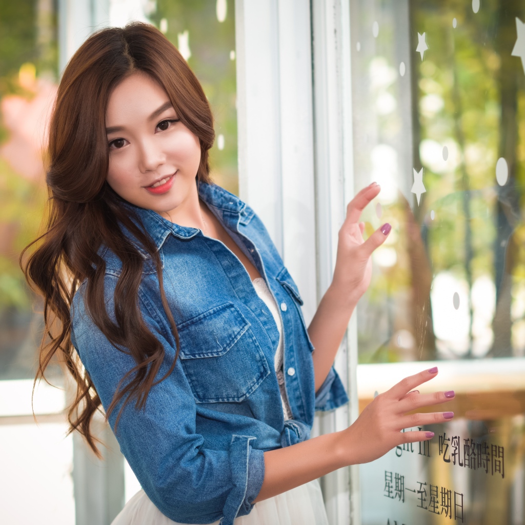 Милая девушка азиатка в джинсовой куртке стоит у окна 