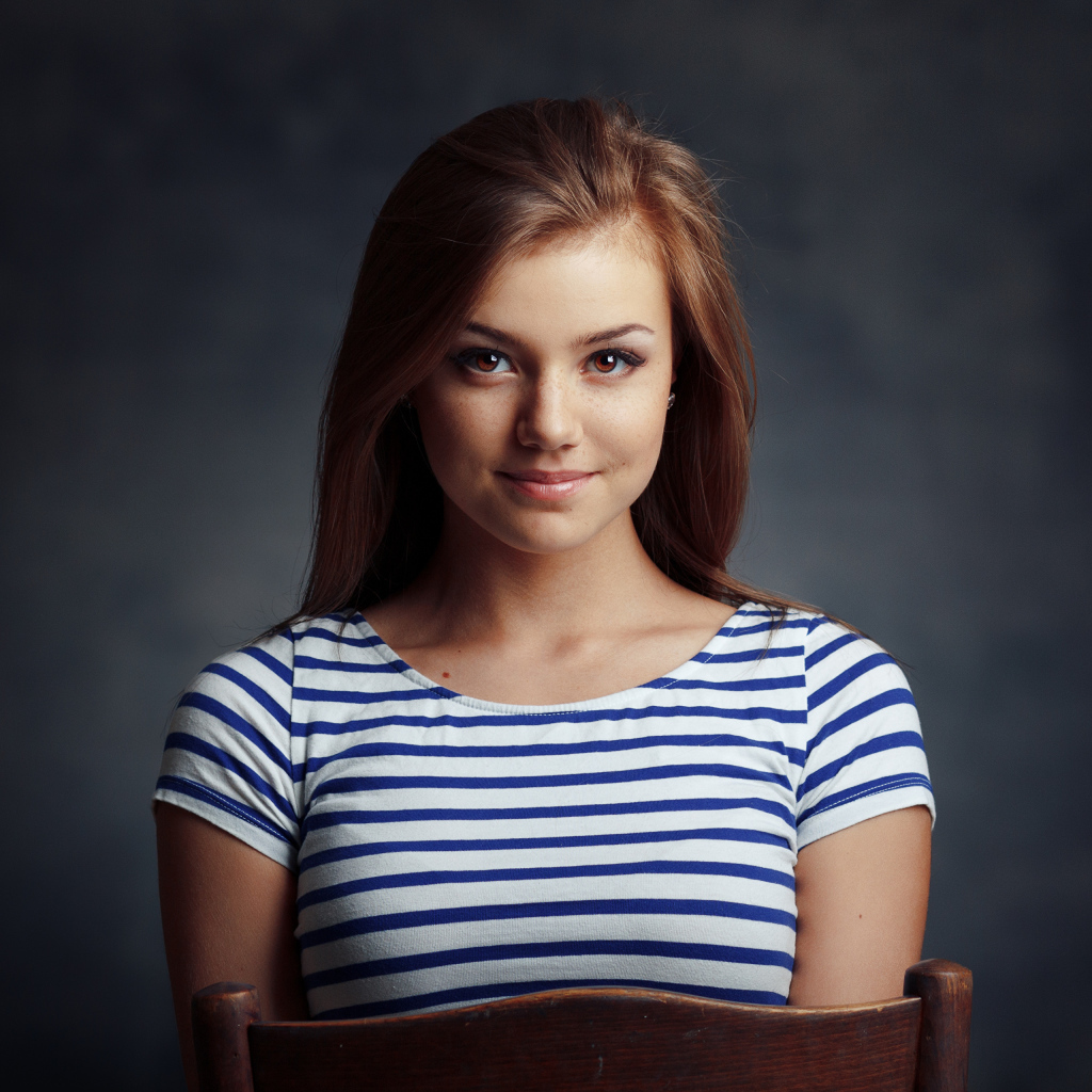 Милая кареглазая девушка в полосатой майке сидит на стуле 