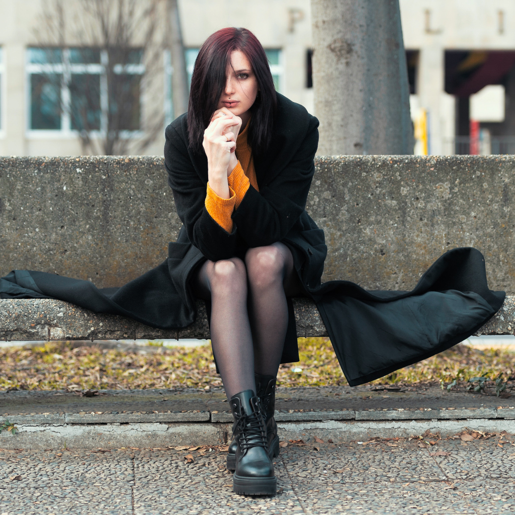 Девушка в черном пальто сидит на бетонной лавке 