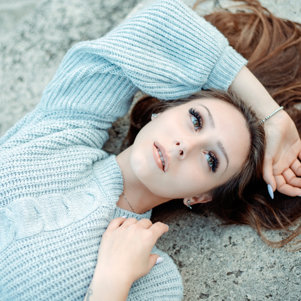 Длинноволосая девушка в голубом свитере лежит на земле 