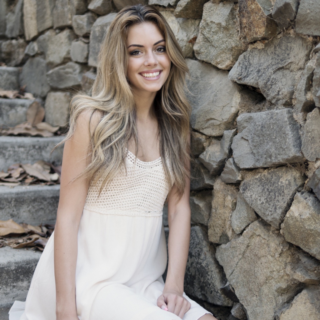 Улыбающаяся  девушка в белом платье стоит на ступеньках у стены