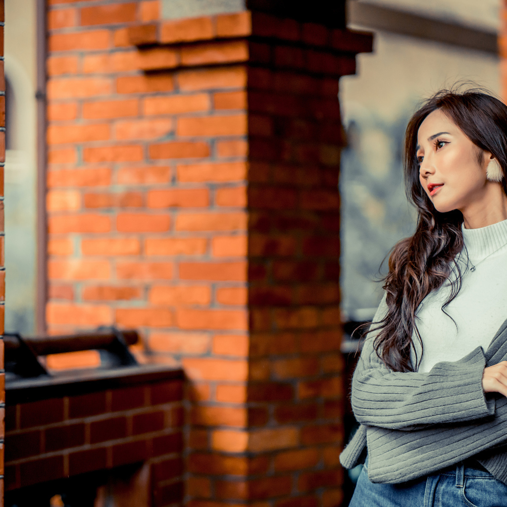 Молодая девушка азиатка в серой кофте у кирпичной стены 