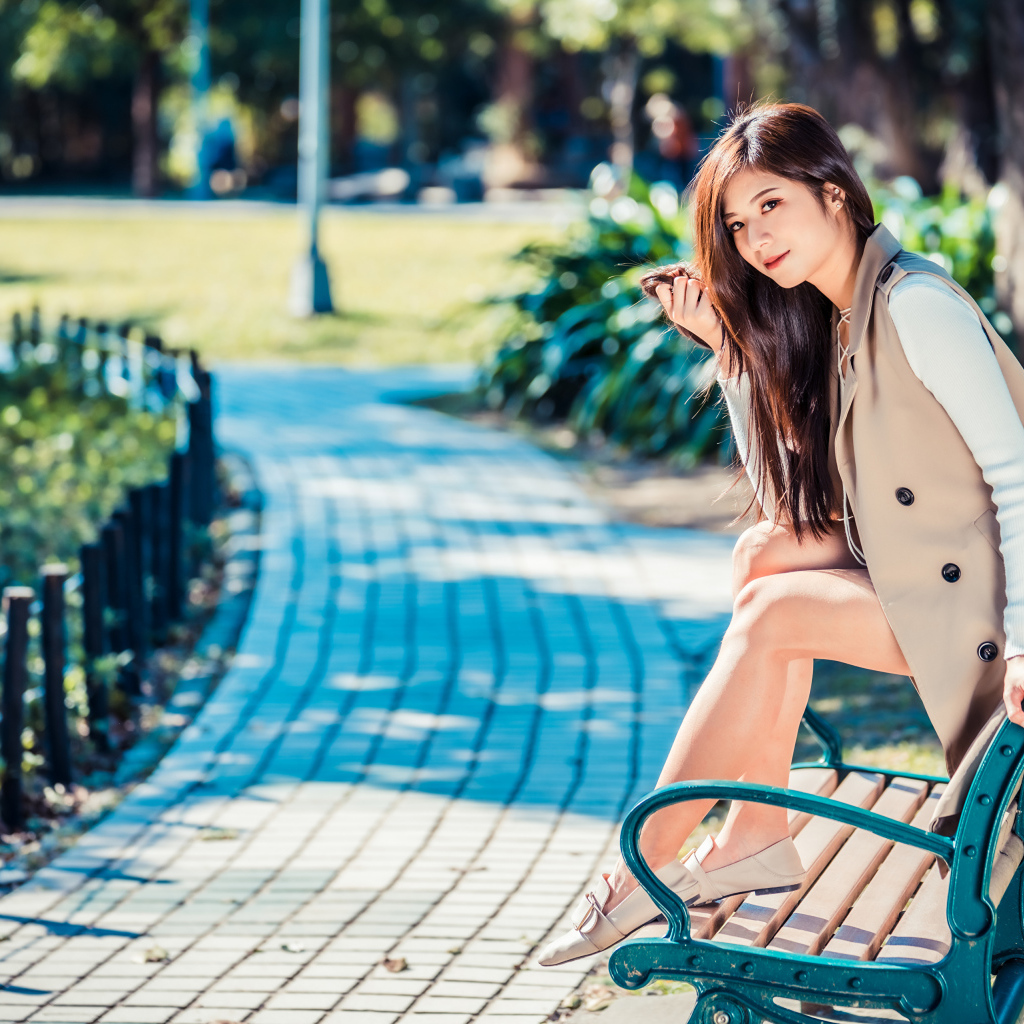 Молодая девушка азиатка сидит в пальто на лавке в парке 