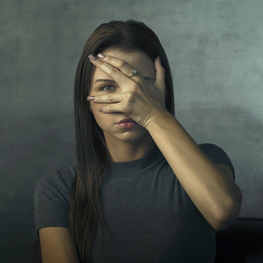 Молодая девушка закрыла лицо рукой на сером фоне