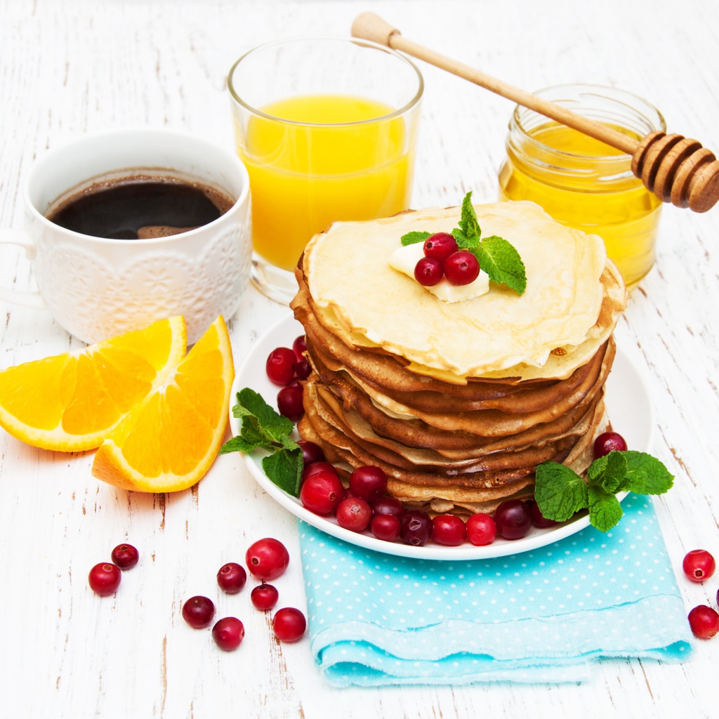 Тонкие румяные блинчики на столе с ягодами клюквы, апельсинами, медом и кофе